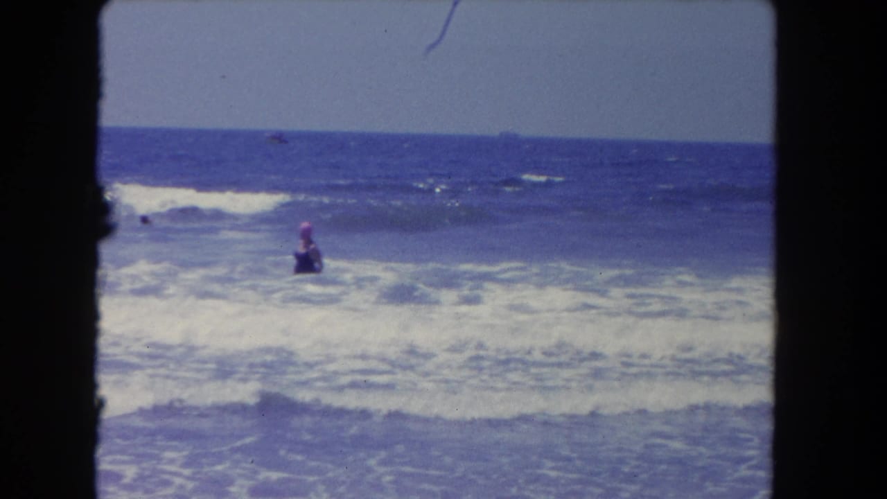1962 Los Angeles beach footage frame with a pink-capped swimmer standing in foamy waves under a clear blue sky.
