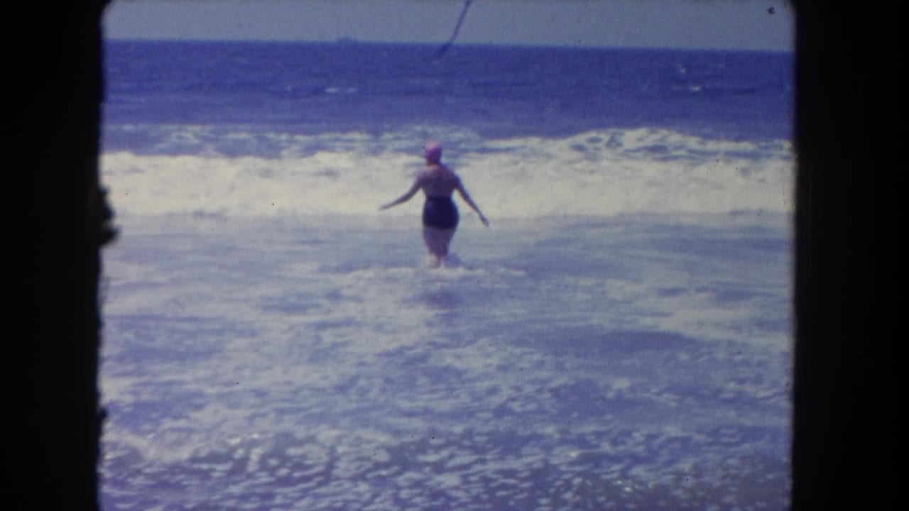 Woman in a dark swimsuit and pink swim cap wading into ocean surf.
