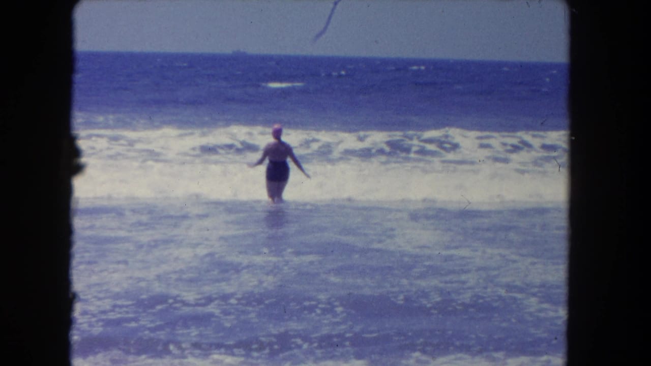 The woman stands farther into the Los Angeles surf, facing the ocean as white waves roll toward shore.