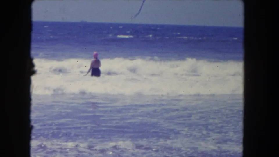 A wave rises around the woman as she remains in the shallow ocean, seen from a handheld 8mm beach viewpoint.