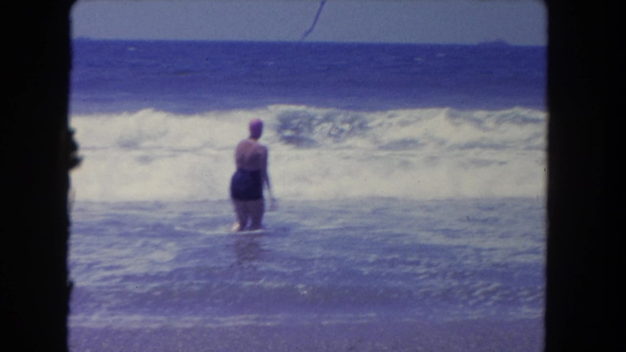 Adult woman in a swim cap stands in shallow ocean surf facing breaking waves.
