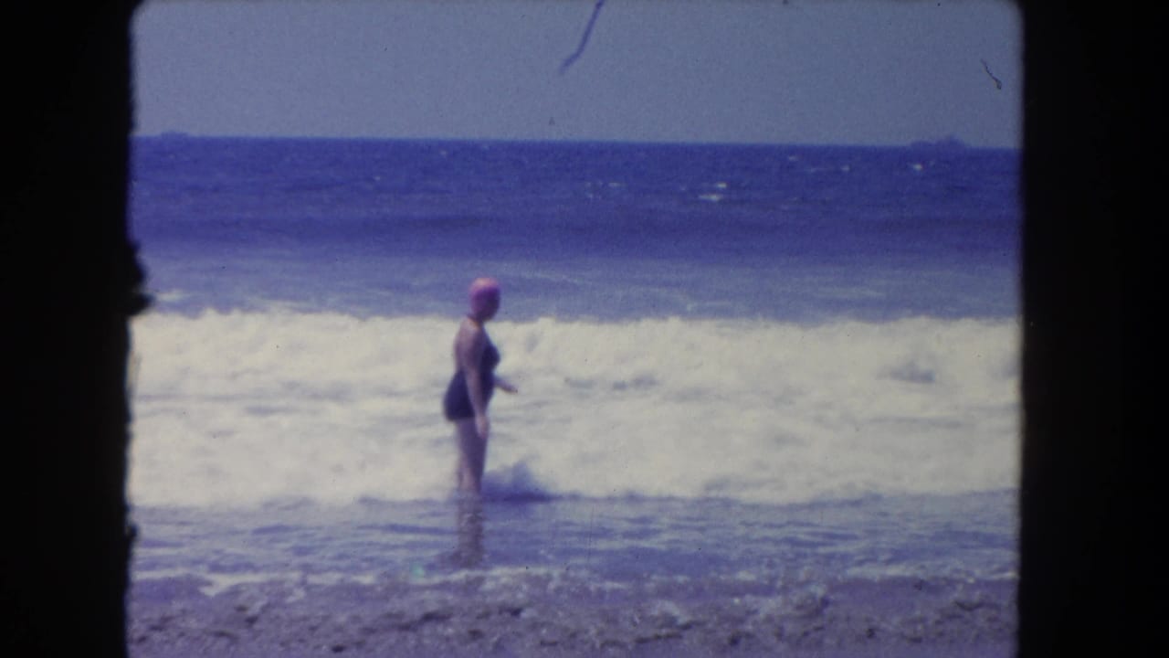 Adult woman in a swim cap stands sideways in foamy ocean water.