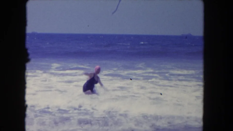 The woman crouches or braces in the shallow surf while a wave breaks around her in this 1962 California beach home movie frame.