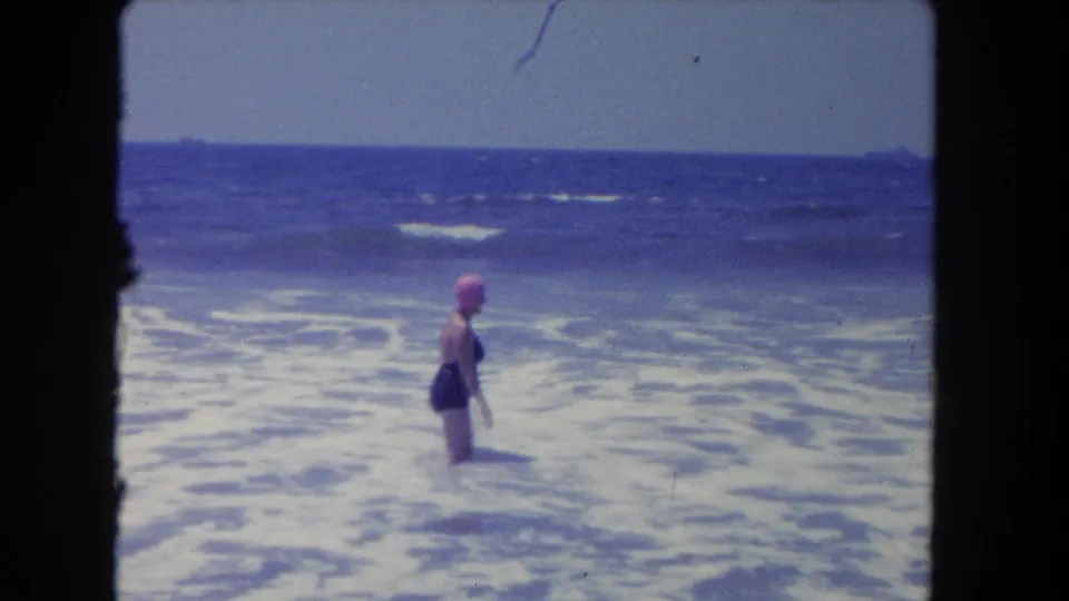 An adult woman in a pink swim cap stands in foamy ocean water at a Los Angeles beach, with open water and surf behind her.