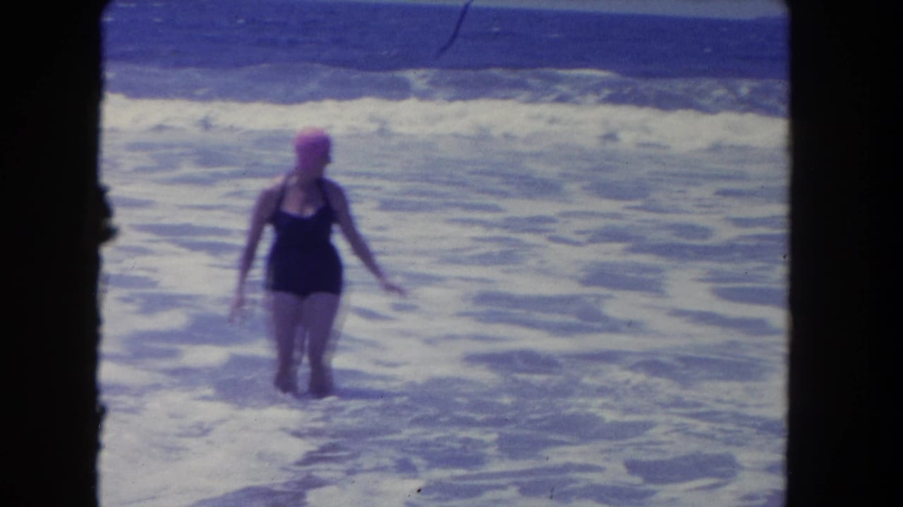 Adult woman in a dark one-piece swimsuit and pink swim cap faces the camera while wading in Los Angeles surf, 1962.