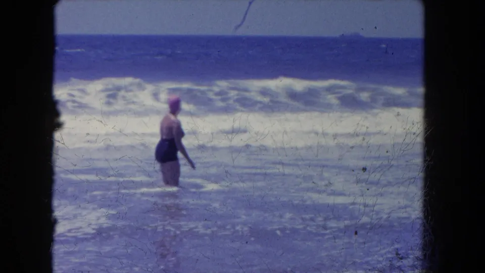 Adult woman in a dark swimsuit and pink cap stands side-on in foamy surf, with blue ocean and horizon behind her.
