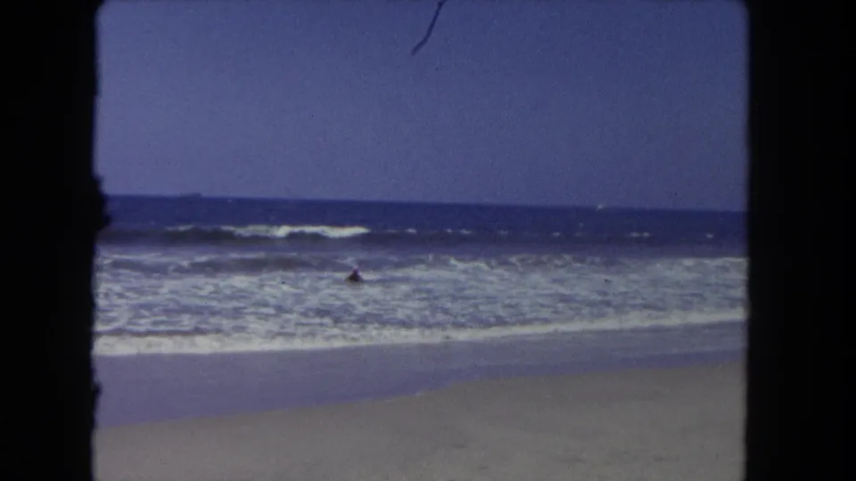 1962 Los Angeles home-movie frame showing ocean surf and wet sand from the beach, with a distant figure in the water.