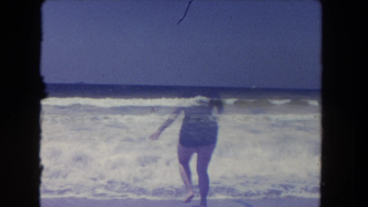 Adult woman in a dark one-piece swimsuit and pink swim cap enters the surf at a Los Angeles beach in 1962.