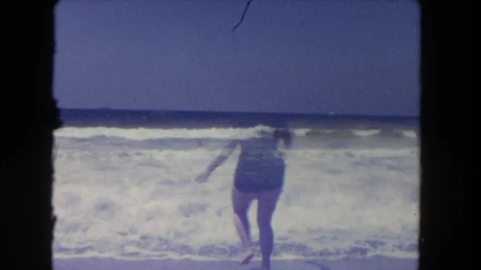 Adult woman in a dark one-piece swimsuit and pink swim cap enters the surf at a Los Angeles beach in 1962.