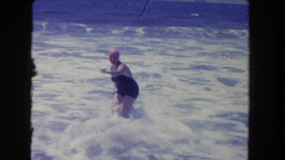 The woman wades through white foam in a dark swimsuit and pink swim cap, filmed close to shore on 8mm.