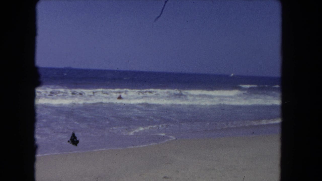 Ocean waves break onto a sandy beach with a distant swimmer and dark film borders.