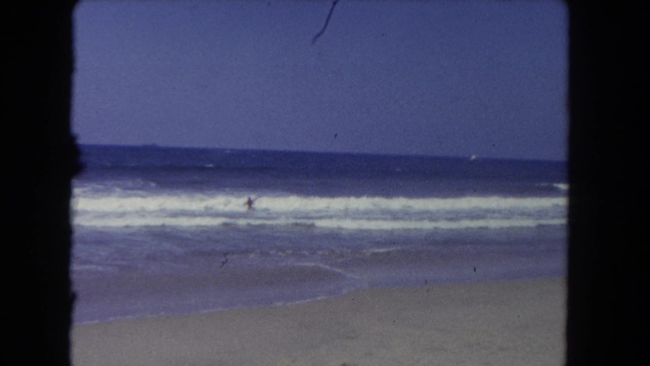 1962 Los Angeles beach footage shows a distant swimmer in choppy surf, with a level horizon, white foam, and sand below.