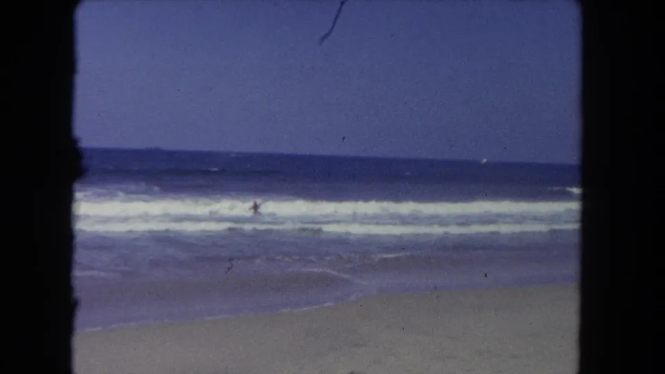 1962 Los Angeles beach footage shows a distant swimmer in choppy surf, with a level horizon, white foam, and sand below.