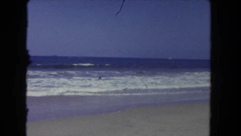 1962 Los Angeles 8mm frame showing foamy ocean waves, sandy beach, open sky, and a small distant swimmer offshore.
