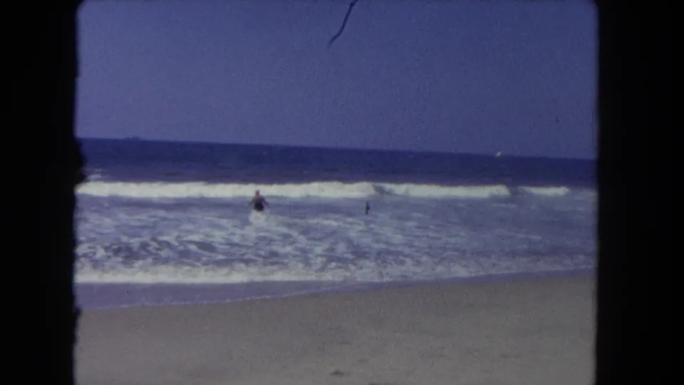 A distant beachgoer appears in the Los Angeles surf, framed by sandy foreground and blue 8mm film color.