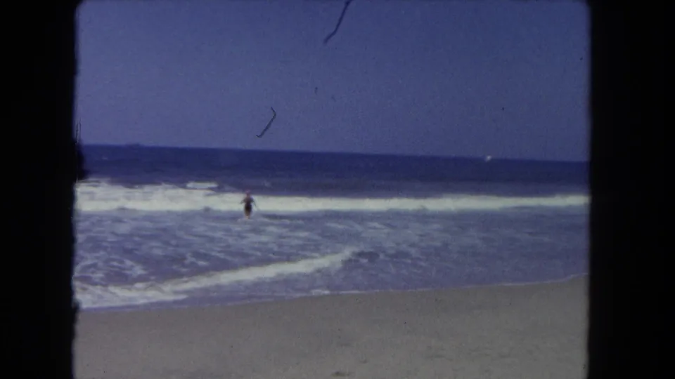 A distant person remains in the shallow surf at a California beach in this soft, grainy 8mm frame.