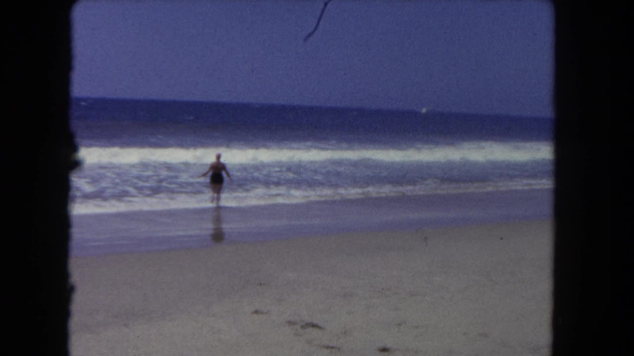 Woman in dark one-piece swimsuit and pink swim cap reaches the shoreline as waves break around her feet, wide open beach and ocean horizon visible, circa 1962.
