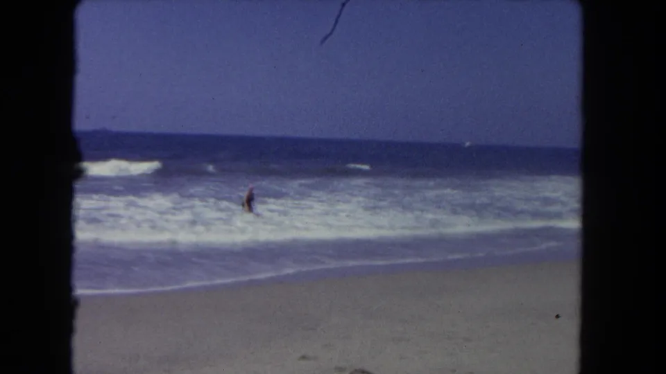 Woman in dark swimsuit and pink swim cap stands deeper in the ocean surf, small figure amid breaking waves, open sandy beach and blue sky, circa 1962.