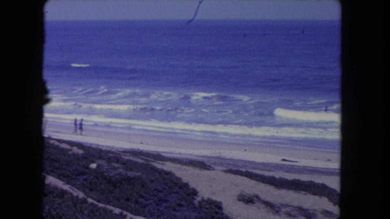 Two distant people stand or walk near the surf on a broad sandy beach.