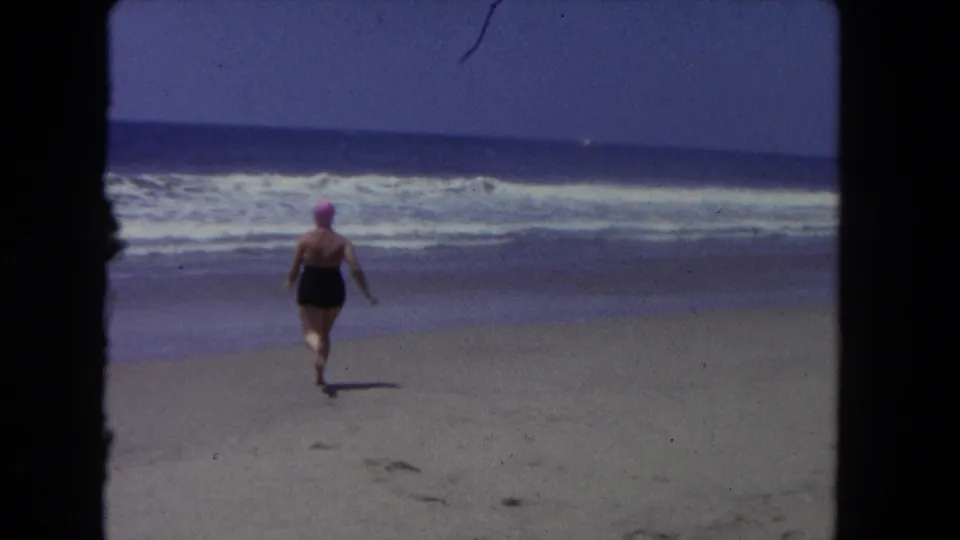 1962 Los Angeles beach scene with a woman in a black bathing suit and pink swim cap running across wet sand toward the breaking surf.