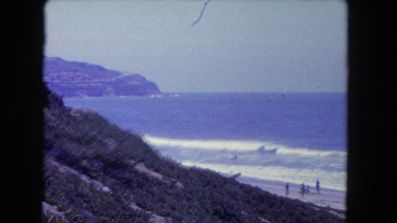 Elevated 8mm film view of a beach — vegetated bluff in foreground, breaking waves, distant rocky headland, tiny figures on sand.