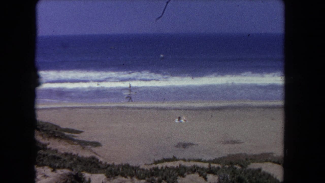 8mm footage, Los Angeles beach ca. 1962: coastal scrub in the foreground, broad sandy shore, breaking surf, open sea, and scattered beachgoers.
