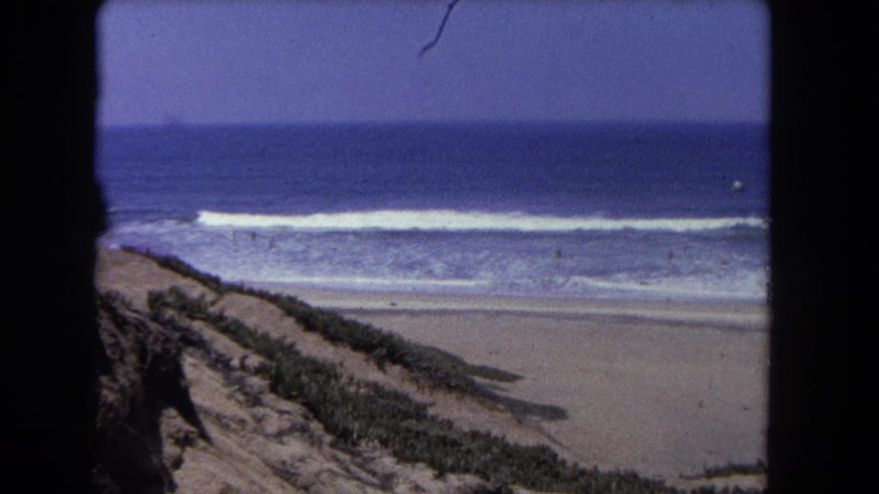 8mm home movie circa 1962, Southern California: same elevated vantage with vegetated dune, wide beach, breaking surf, and blue ocean; slight wave variation from prior frame.