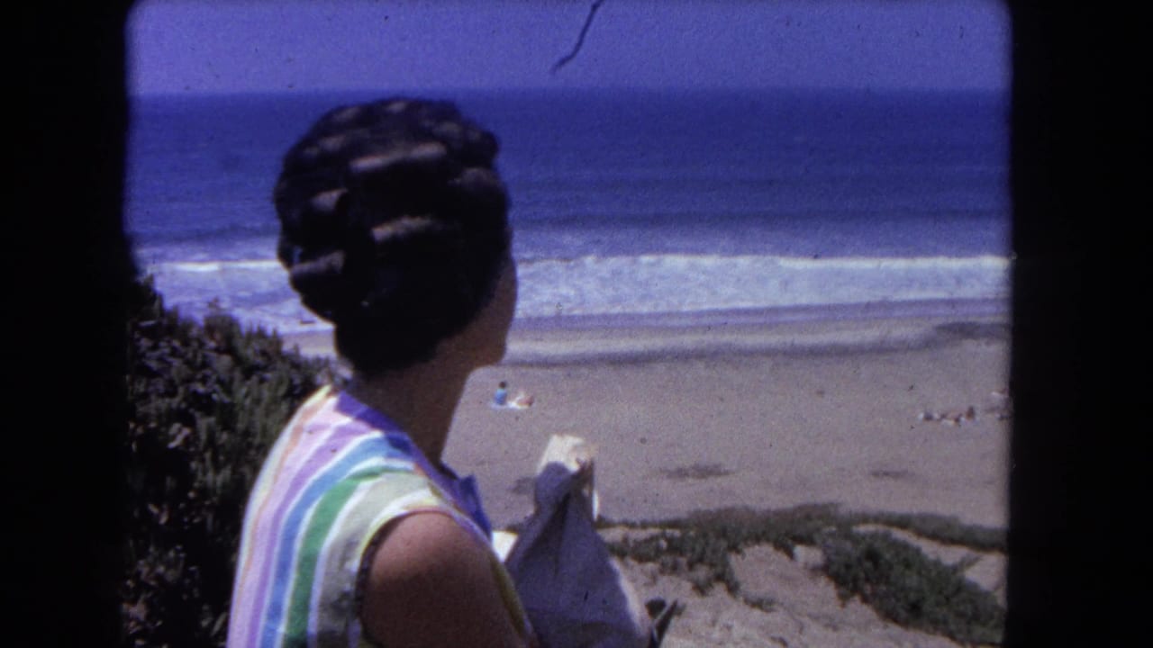 Dark-haired woman in striped blouse, rear view, overlooking California coastline; 8mm home movie framing, circa 1962.