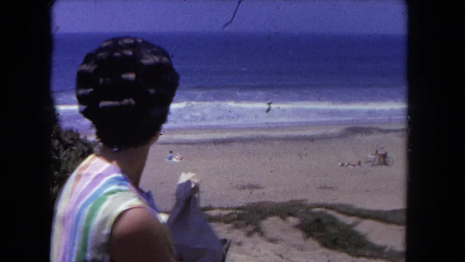 Wider beach view with woman in striped blouse in foreground; distant beachgoers visible on the sand; 8mm home movie, Los Angeles, circa 1962.