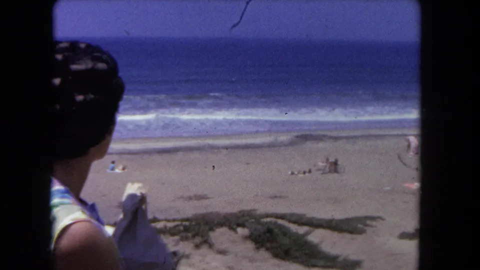 Close rear view of woman in striped blouse; broad beach and ocean horizon behind her; 8mm home movie, California, circa 1962.