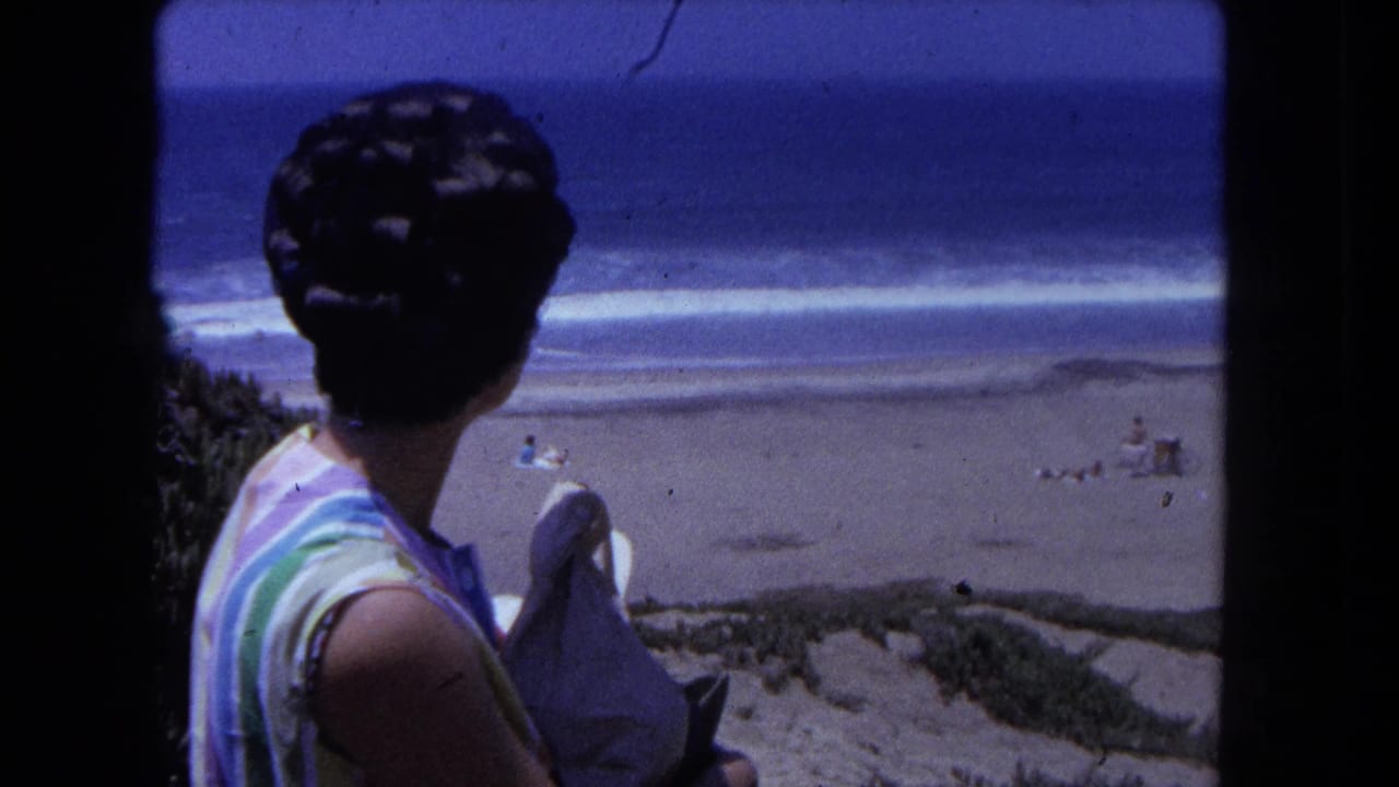 Woman in horizontal-stripe sleeveless top with 1960s-era updo hairstyle at an elevated dune edge overlooking a sandy beach and ocean, circa 1962.