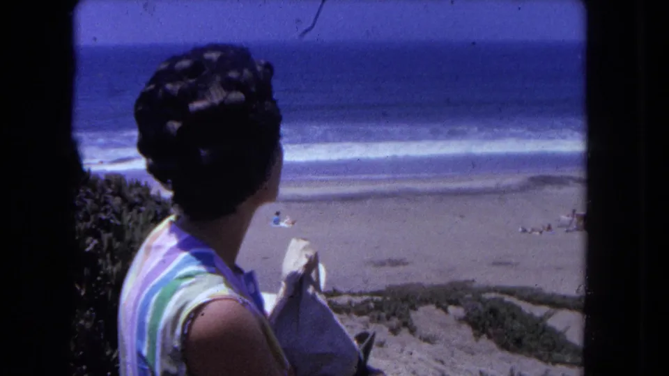 Woman in horizontal-stripe sleeveless top partially in profile holding a white item; sandy beach with breaking waves and coastal vegetation visible below, circa 1962.
