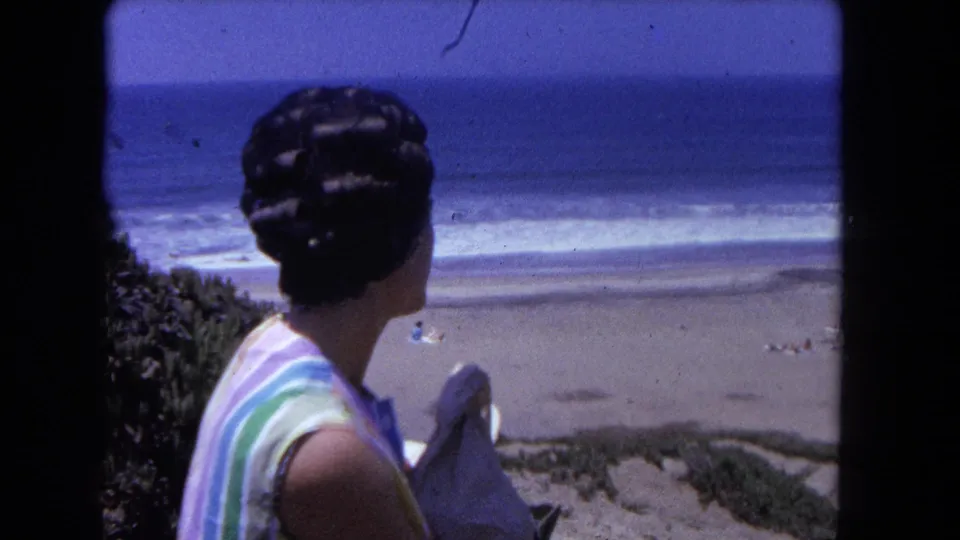 Rear view of woman in horizontal-stripe sleeveless top with vintage updo, standing at a coastal dune overlook with sandy beach and ocean waves below, circa 1962.