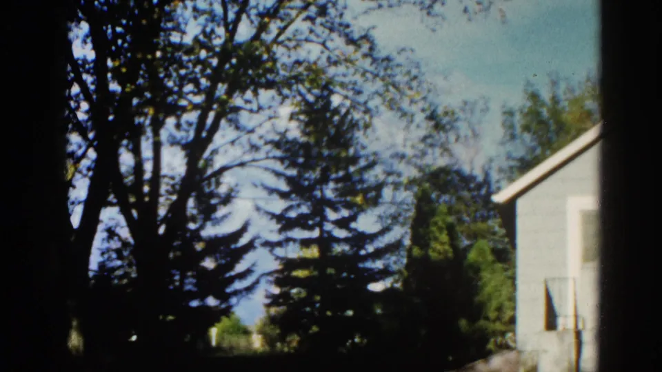 8mm home movie frame, circa 1962, Illinois — same backyard treetop composition with residential house corner and blue sky, window or vent detail at right.