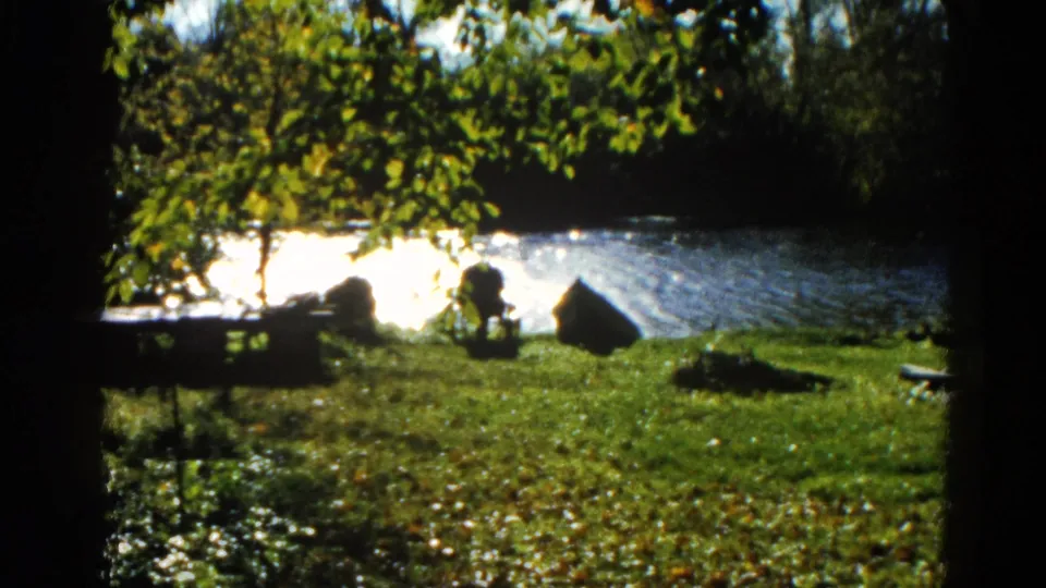 8mm home movie frame, circa 1962, Illinois — suburban backyard with green lawn, pond reflecting bright sunlight, surrounding trees, and residential house corner.