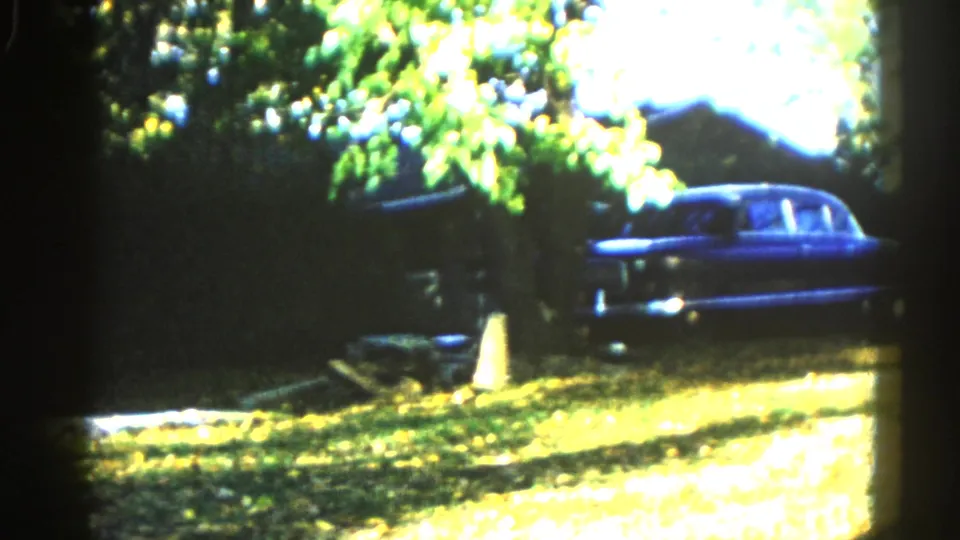 Large dark-blue American sedan parked in a leaf-dappled suburban yard under a deciduous canopy; Illinois home movie, circa 1962, 8mm color.