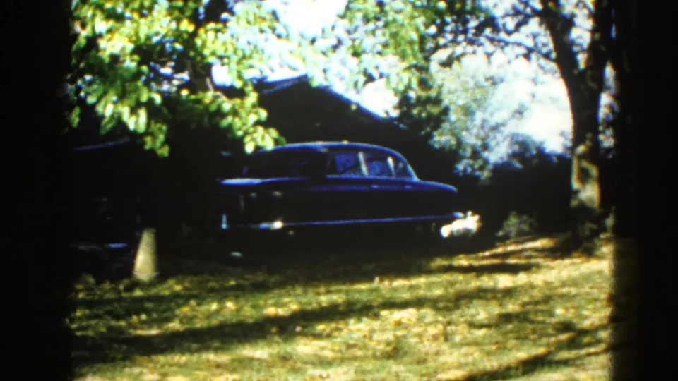 Large dark-blue American sedan parked under a leafy tree canopy in a suburban yard; 8mm home movie, Illinois, c. 1962.