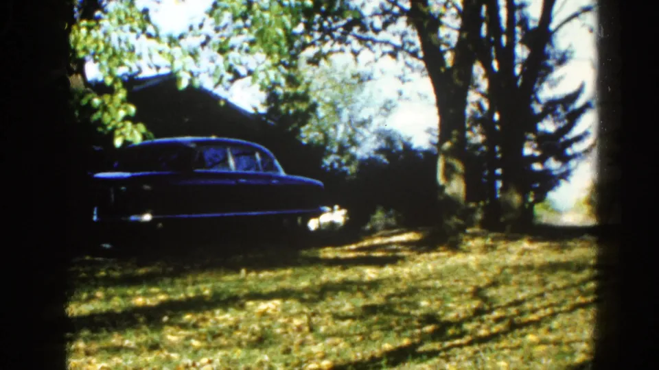 Mature deciduous trees dominate the frame; a dark-blue sedan is partially visible at left in a sunlit suburban Illinois yard, c. 1962, 8mm home movie.