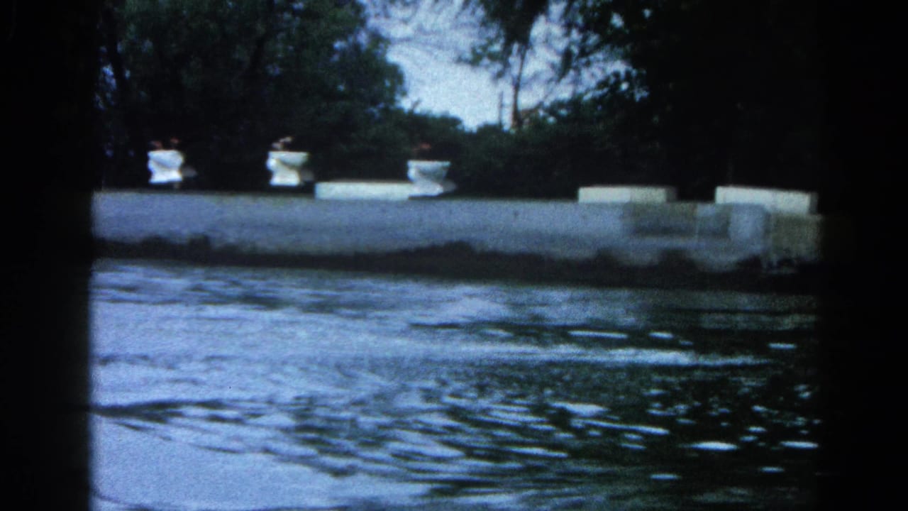 Calm lake in foreground with a concrete dock and white bollards at midframe; dense tree line behind; 8mm film vignetting on edges.