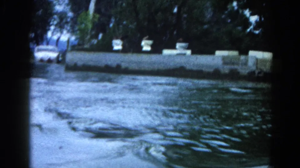 8mm home-movie frame, Illinois 1962: concrete dock edge with bollards, a light-colored boat hull at frame left, and indistinct figures near the waterline.