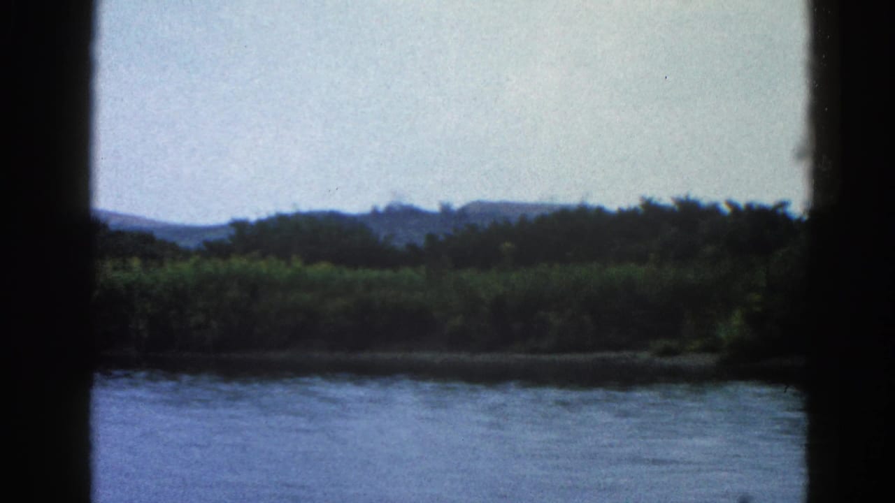 Continued river view with dense far-bank treeline and low hills under a pale sky, similar framing to opening frame.