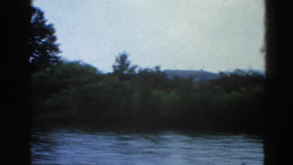 8mm home movie frame, circa 1962 Illinois — closer riverbank with trees at left, open water foreground, low hills in background.