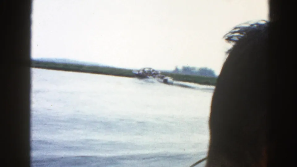 8mm home movie frame, circa 1962 Illinois — silhouetted passenger head in foreground, diagonal boat wake or tow activity across frame, pale overexposed sky and distant treeline.