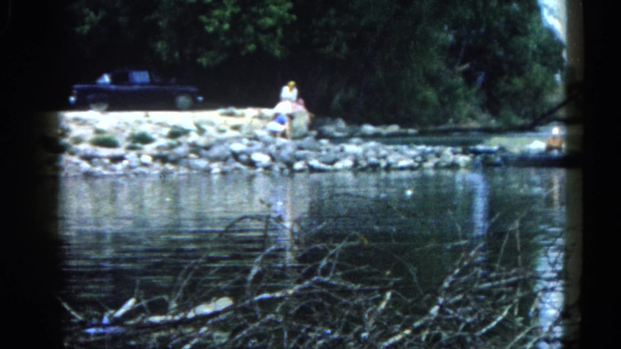 8mm home movie frame of a figure seated on a gravel riverbank in Illinois, c. 1962, with a large dark American car parked on the shore behind.