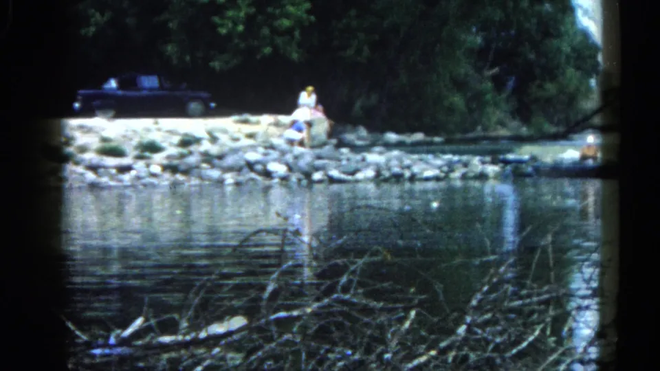 8mm home movie frame of a figure seated on a gravel riverbank in Illinois, c. 1962, with a large dark American car parked on the shore behind.