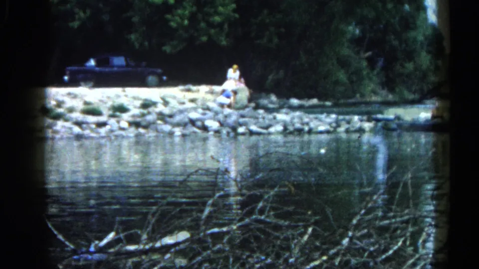 8mm shot across a reflective Illinois river to a stone shore where a figure sits near a dark vintage automobile beneath dense summer foliage, c. 1962.