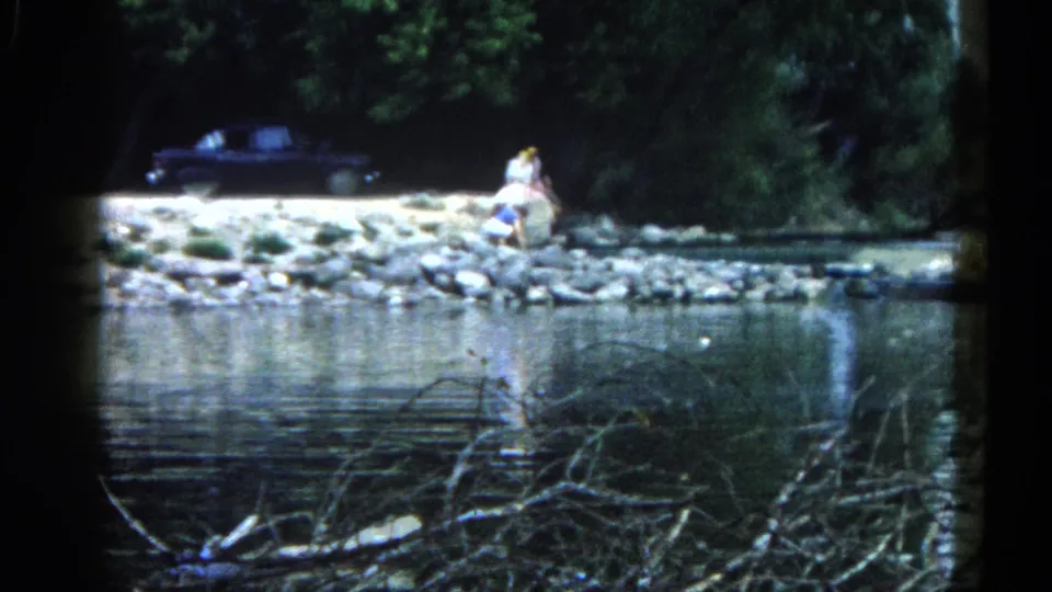 Final frame of 8mm Illinois riverside clip, c. 1962; seated figure and dark American car on rocky shore with summer trees and calm water throughout.