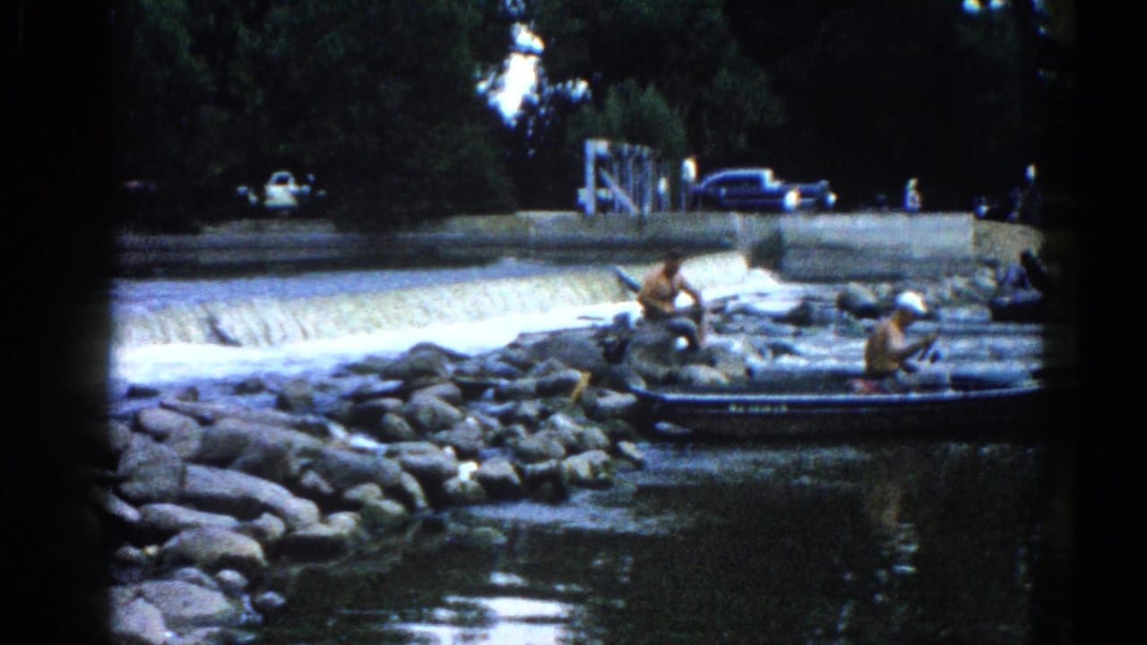 8mm home movie frame, c.1962 Illinois — adults at a small boat on a boulder-lined shore; a low concrete dam weir and vintage parked cars are visible behind.