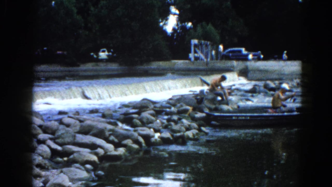 Adults near a small boat on a boulder shoreline, with a concrete weir spilling water in the background.