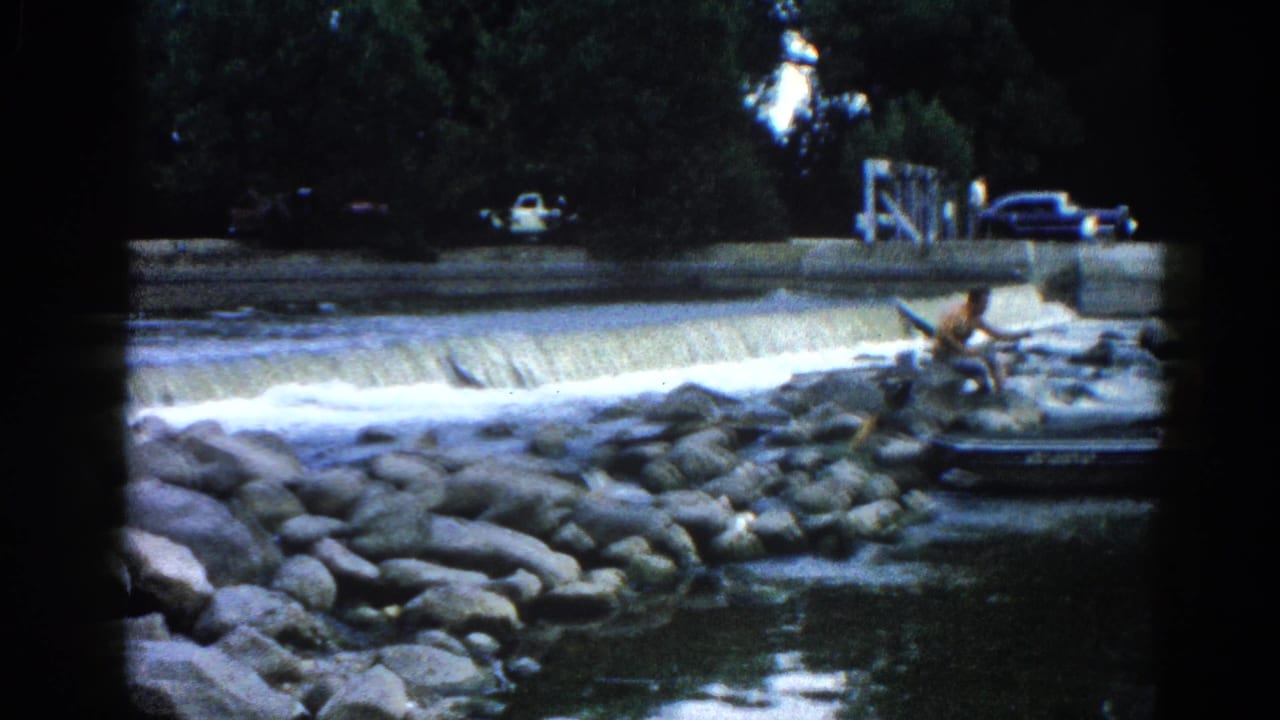 8mm home movie frame, c.1962 Illinois — wider angle reveals concrete weir, small dark boat on boulder shore, and dark-bodied vintage cars parked under a dense tree canopy.